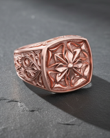 Ornate men's Maltese Cross ring in rose gold, showcasing the detailed floral motif and stippled texture on a slate surface.