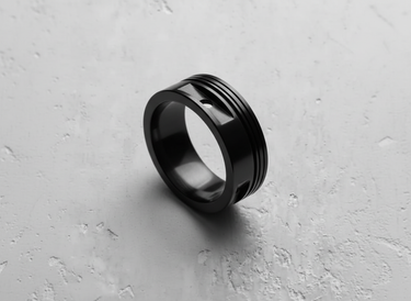 Black rhodium plated silver piston ring showing recessed grooves and wrist pin cutout on a textured white surface.