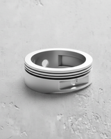 Silver piston ring with dual recessed grooves and piston pin cutout detail, shown on a textured gray surface.