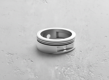 Silver piston ring with dual recessed grooves and piston pin cutout detail, shown on a textured gray surface.