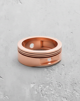 Rose gold plated silver piston ring showcasing the recessed compression grooves and gudgeon pin cutout on a grey surface.