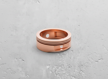 Rose gold plated silver piston ring showcasing the recessed compression grooves and gudgeon pin cutout on a grey surface.