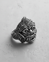 Silver Balinese mask ring showing the ornate headdress and wide-eyed, fanged expression on a textured light gray surface.