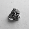 Silver Balinese mask ring showing the ornate headdress and wide-eyed, fanged expression on a textured light gray surface.