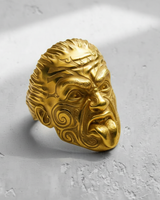 Gold vermeil Maori Warrior ring with fierce expression and tribal carvings, shown at an angle on a textured surface.