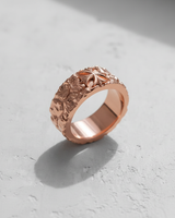 Rose gold plated silver cross ring showcasing its rough, organic band texture and stylized floral cross on a light surface.