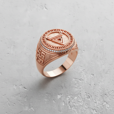 Rose gold plated geometric symbol ring with an inverted triangle, sunburst border, and pavé stones on a textured surface.