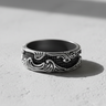 Men's silver wave grid ring with ornate flowing carvings and a central square pattern, shown at an angle on a textured surface.