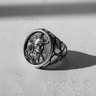 Oxidized Silver Rooster Cameo Ring with a mythological warrior profile, shown at an angle on a textured white surface.