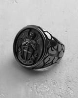 Silver Saint Michael ring showing the detailed archangel relief with shield, sword, and wings on a textured white surface.
