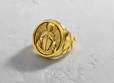Gold vermeil Saint Michael ring on a textured white surface, showcasing the detailed Archangel, shield, and textured band.