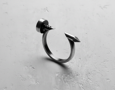 Oxidized Silver Nail Ring with a flat head and pointed tip shown on a textured, light gray concrete surface.