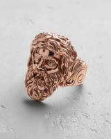 Rose gold Zeus ring from a side profile, displaying the flowing hair and band design against a light gray textured background.