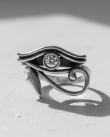 Silver Eye of Horus ring with a central gemstone, shot in black and white on a textured white surface casting a shadow.