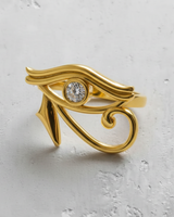 Gold-plated Eye of Horus ring with a sparkling central diamond, photographed from above on a light grey textured background.