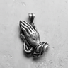 Silver praying hands pendant with detailed fingers and cuffs, shown flat on a textured light gray surface.