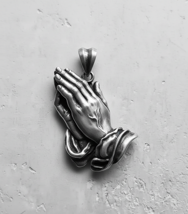 Silver praying hands pendant with detailed fingers and cuffs, shown flat on a textured light gray surface.