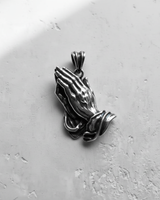 Angled view of the silver praying hands pendant, with shadows highlighting the oxidized finish and sculptural depth.