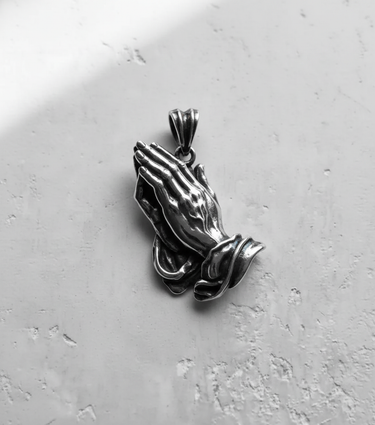 Angled view of the silver praying hands pendant, with shadows highlighting the oxidized finish and sculptural depth.