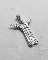 Silver Jesus Christ pendant with outstretched arms, showing detailed religious scenes carved into the robe, on a textured grey surface.