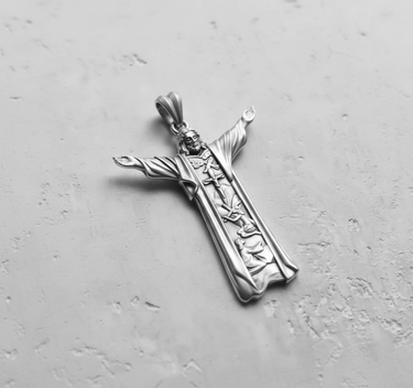 Silver Jesus Christ pendant with outstretched arms, showing detailed religious scenes carved into the robe, on a textured grey surface.
