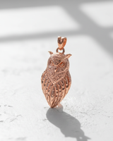Rose gold plated silver owl pendant standing upright, catching the light to show warm tones on a white textured surface.