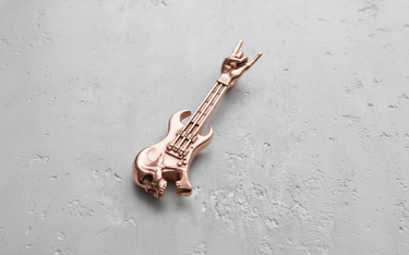 Rose gold-plated silver guitar skull pendant, angled to highlight the fretboard and rock-hand detail against a concrete background.