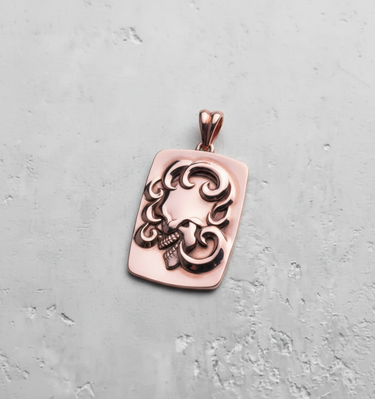 Rose gold ornate skull pendant featuring a side-profile skull and swirling patterns, shot from a top-down angle.
