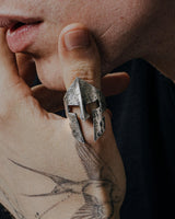 A man's hand with a tattoo wearing a sterling silver Spartan helmet ring with a rugged, oxidized, battle-worn finish.