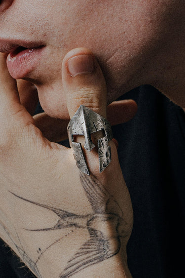 A man's hand with a tattoo wearing a sterling silver Spartan helmet ring with a rugged, oxidized, battle-worn finish.