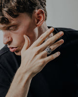 Close-up of a man's hand touching his face, highlighting the handcrafted silver owl ring worn as a statement piece of jewelry.