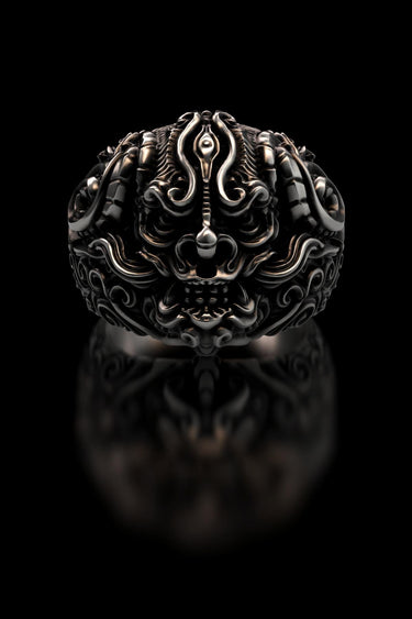 Sterling silver Oni mask ring with intricate carvings and oxidized finish shown in a studio shot on a reflective black background.