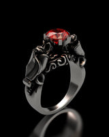 Silver bat ring with a round-cut red stone, showing the detailed gothic design of two bats holding the gem on a black background.