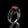 Silver bat ring with a round-cut red stone, showing the detailed gothic design of two bats holding the gem on a black background.