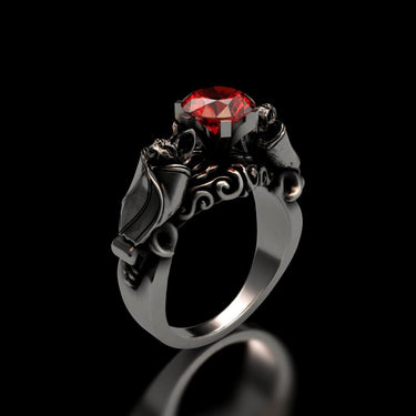 Silver bat ring with a round-cut red stone, showing the detailed gothic design of two bats holding the gem on a black background.