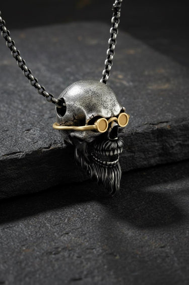 Silver bearded skull pendant with gold vermeil goggles shown at an angle on a dark, textured slate surface for a dramatic look.