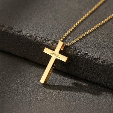 Personalized cross necklace in 18k gold vermeil with the name 'Jessica' engraved, resting on a dark textured stone.