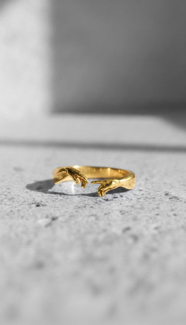 Gold-plated version of the Creation of Adam Ring, showcasing the iconic two hands design on a textured gray surface.
