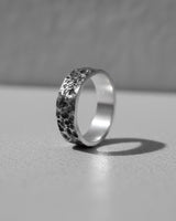 Black and white photo of a Moon Surface Silver Ring with a heavily cratered, hammered texture, standing on a light surface.