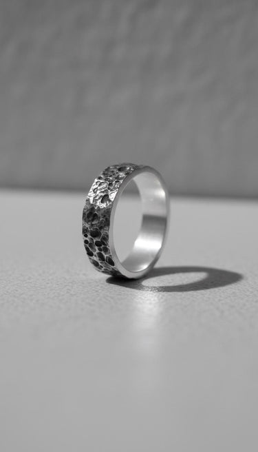 Black and white photo of a Moon Surface Silver Ring with a heavily cratered, hammered texture, standing on a light surface.