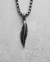 Close-up of a silver feather pendant with detailed carving, hanging from a sterling silver box chain on a textured white background.