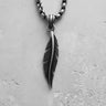 Close-up of a silver feather pendant with detailed carving, hanging from a sterling silver box chain on a textured white background.