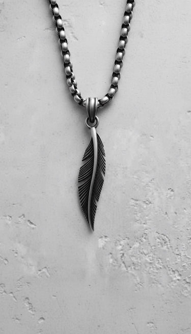 Close-up of a silver feather pendant with detailed carving, hanging from a sterling silver box chain on a textured white background.