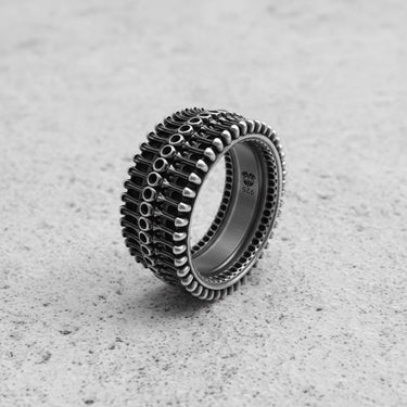 Silver biomechanical ring with three rows of industrial-style detailing, shown at an angle on a textured concrete surface.