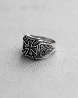 Silver Iron Cross ring with oxidized finish and detailed oak leaf carvings on the band, shown on a textured white surface.
