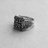 Silver Iron Cross ring with oxidized finish and detailed oak leaf carvings on the band, shown on a textured white surface.