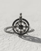 Handmade silver compass pendant with North Star design and cardinal points, lying flat on a textured white surface.