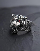 Men's silver tiger ring with detailed oxidized stripes and red gemstone eyes, shown at an angle on a dark slate surface.