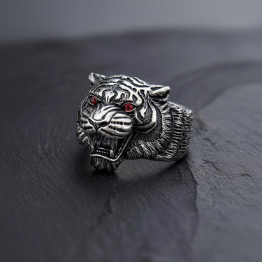 Men's silver tiger ring with detailed oxidized stripes and red gemstone eyes, shown at an angle on a dark slate surface.