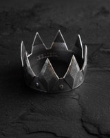 Oxidized silver crown ring with a textured geometric design and studded band, displayed on a dark, cracked slate surface.